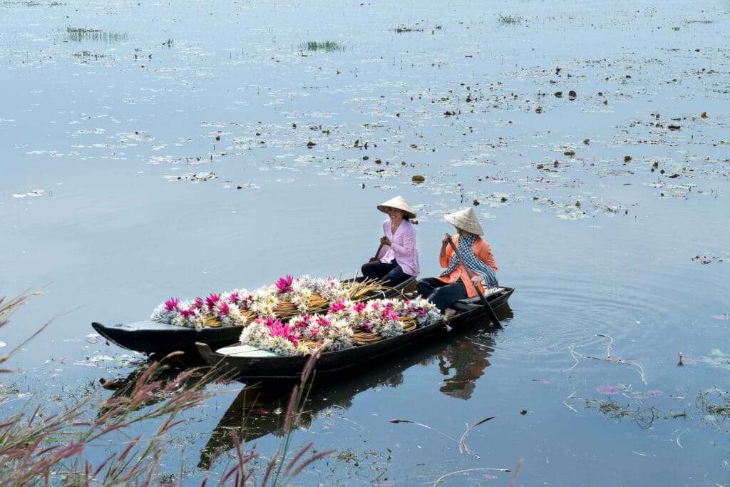 Two women paddle a canoe filled with vibrant flowers on a serene lake. Peaceful outdoor scene.