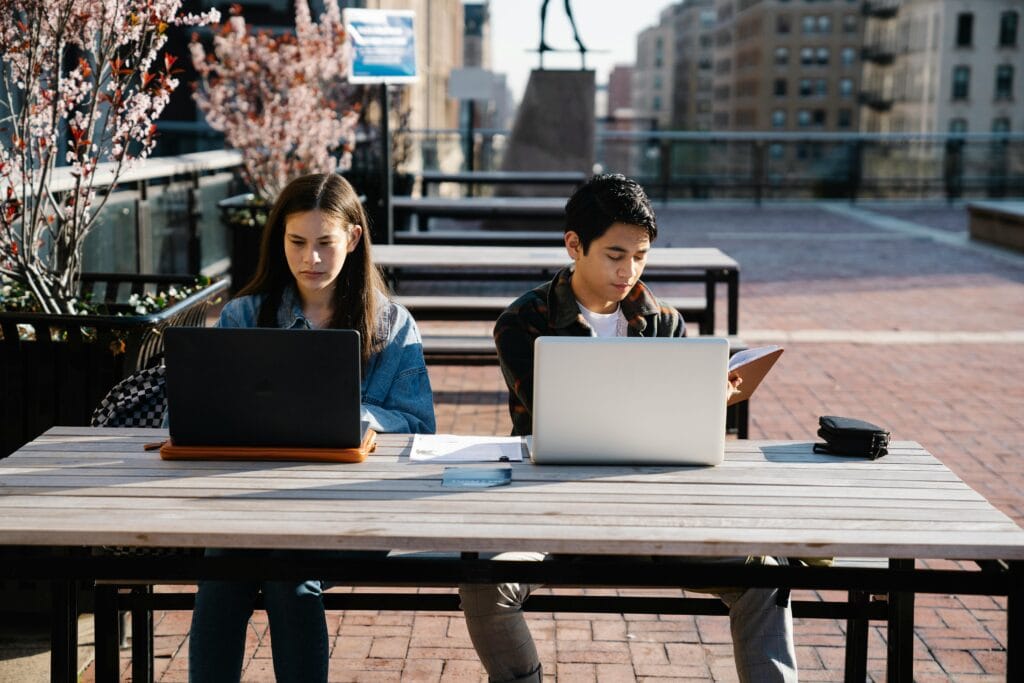 Young adults working on laptops outdoors at a table in a city setting, focused and concentrated.