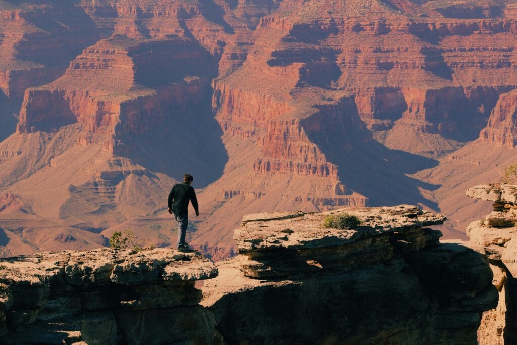 A man hikes along the rocky edge of the Grand Canyon under a clear blue sky, capturing the essence of outdoor adventure.